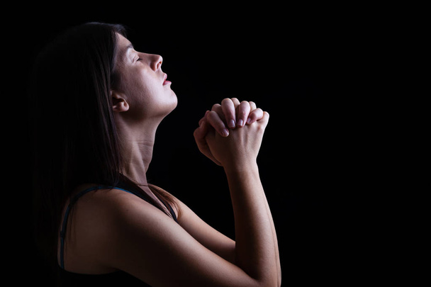 Acercamiento de una mujer fiel orando, las manos dobladas en adoración, la cabeza hacia arriba y los ojos cerrados en fervor religioso, sobre un fondo negro. Concepto de religión, fe, oración y espiritualidad
. - Foto, imagen