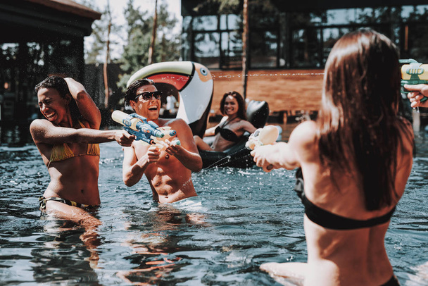 Group of Young Smiling Friends having Fun in Pool. Young Happy People Playing Together with Colorful Water guns in Outdoor Swimming Pool. Friends at Pool Party. Summer Vacation Concept - Fotó, kép