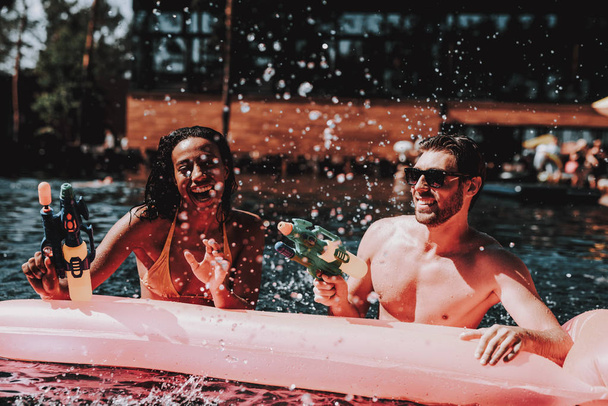 Young Smiling Couple having Fun in Swimming Pool. Young Happy People Playing Together with Colorful Water guns in Outdoor Swimming Pool. Friends at Pool Party. Summer Vacation Concept - Zdjęcie, obraz