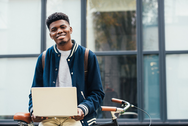 smiling african american freelancer working with laptop while leaning on bike - Photo, Image