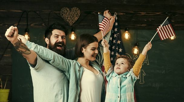 Kid with parents in classroom with usa flags, chalkboard on background. American cheerful family with son and usa flags. Patriotic education concept. Parents teaching son american traditions playing. - Foto, immagini