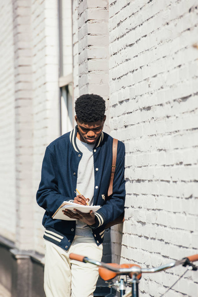 african american freelancer writing documents and standing near white brick wall - Foto, afbeelding