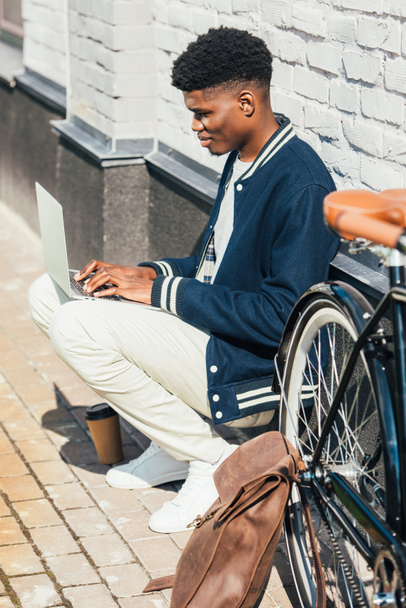 freelancer afro-americano elegante trabalhando com laptop perto de bicicleta com mochila de couro na cidade
 - Foto, Imagem