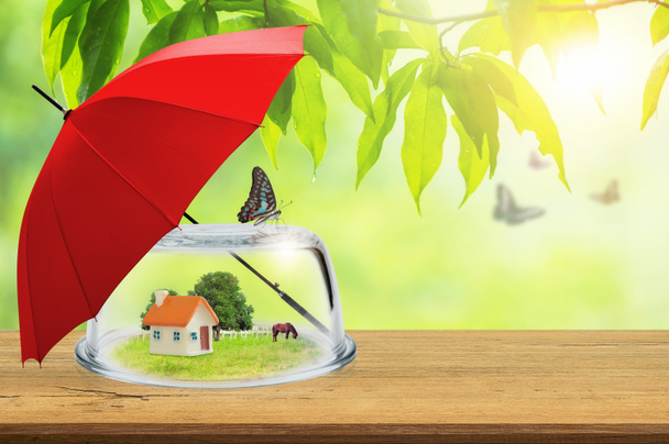 Sachversicherungen, verschwommen Spielzeug Haus in Glaskuppel auf Holzböden und einem roten Regenschirm gegen natürliche auf Hintergrund, Konzept der Versicherung. - Foto, Bild