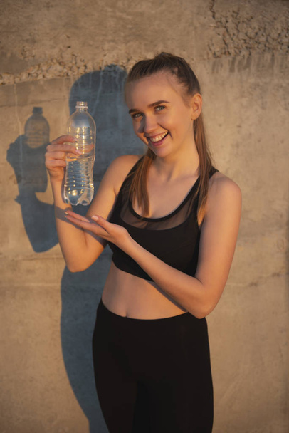 Portrait of beautiful caucasian smiling girl after training with bottle of water. Dressed in black leggings and shirt. Stone background on sunset. - Foto, immagini