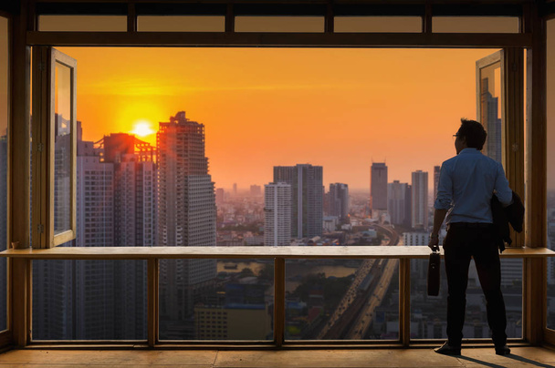 Concept vision, Young businessman wearing comfortable casual suit jacket standing holding a work bag in Working room near the glass window and looking through the modern buildings in the skyline. - Foto, Bild