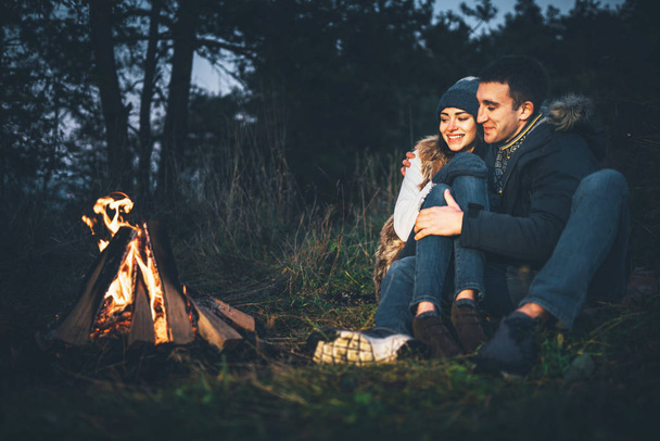 Pretty young couple relaxing near bonfire in the forest at evening time - Фото, изображение