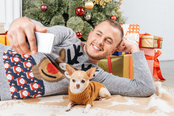 Glimlachende man nemen selfie met chihuahua hond met kerstboom op achtergrond thuis - Foto, afbeelding