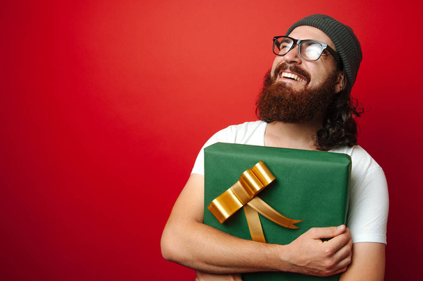 Portrét člověka šťastný veselý vousatý bokovky drží velký dárek na Vánoce / Nový rok nad červeným pozadím s volného místa pro text - Fotografie, Obrázek