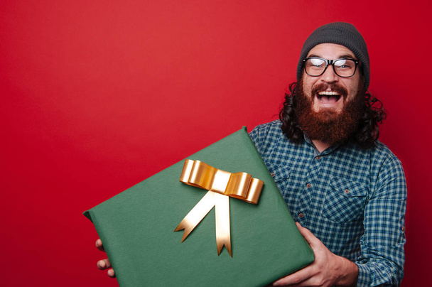 Портрет изумленного бородатого мужчины с большой подарочной коробкой на красном фоне
 - Фото, изображение