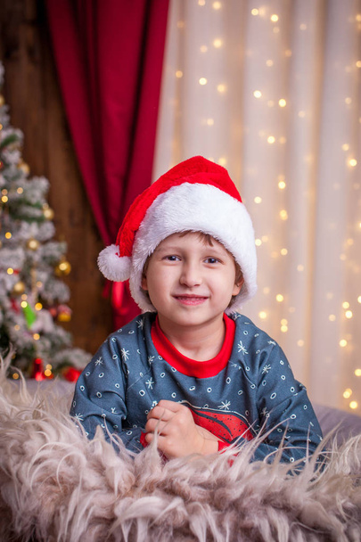 boy is smiling watching the Christmas tree in the lights and the window with the garland surrounds the holiday atmosphere - Zdjęcie, obraz