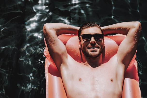 Young Smiling Man in Sunglasses on Air Mattress. Handsome Bearded Man wearing Sunglasses Relaxing by Lying on Inflatable mattress in Outdoor Swimming Pool. Summer Vacation Concept - Foto, afbeelding