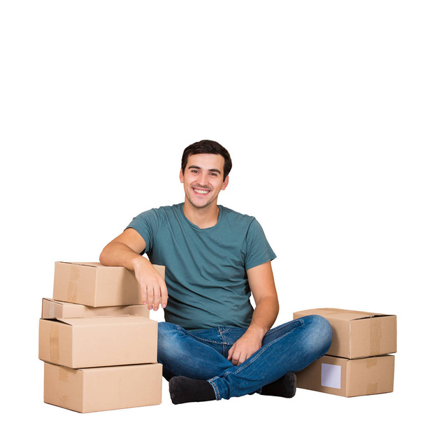 Mladá vzrušený muž sedící na podlaze libové na lepenkové krabice připravené na stěhování do nového domu, jeho věci a zbytek izolované na bílém pozadí. - Fotografie, Obrázek