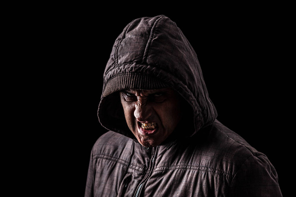 Very angry and aggressive man hiding in the shadows, with the face partly hidden with the hood, and standing in the darkness. Low key, black background. Concept for anger, rage, violence, danger - Photo, Image
