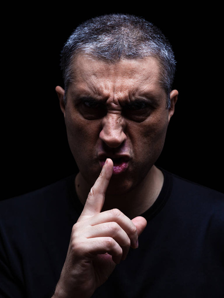 Furious mature man with an aggressive look making the silence sign in a violent and threatening way. Low key, black background. Concept for threat, anger, rage, violence, danger, menace, fury.  - 写真・画像