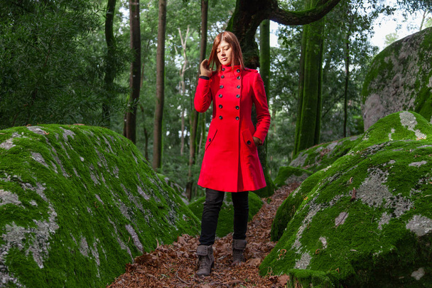 Νεαρή γυναίκα λυπημένοι μόνο το περπάτημα στο δάσος μονοπάτι φορώντας κόκκινο μακρύ παλτό ή πανωφόρι. Κορίτσι περνάει κομμάτι για βόλτα στο δάσος της φύση πάρκο κατά τη διάρκεια της πτώσης, το φθινόπωρο ή το χειμώνα - Φωτογραφία, εικόνα