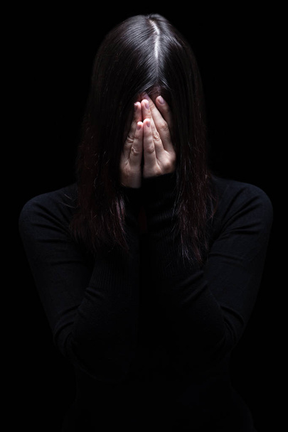 Emotional woman crying and covering the face with the hands hiding the tears, on a black or dark background. Concept for victim, depression, pain, grief, mourn, despair, sadness, fear. - Photo, Image