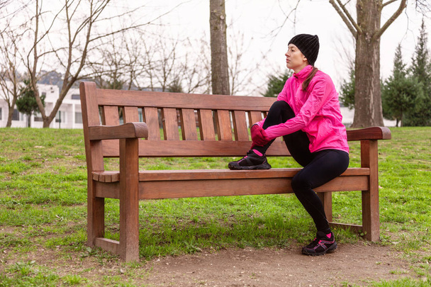 Kadın atlet yorgun veya bir kentsel park eğitim yolda bir soğuk kış gününde bir bankta oturan depresif. Giymiş pembe rüzgarlık, bere, eldiven ve tayt çalışan genç kadın - Fotoğraf, Görsel
