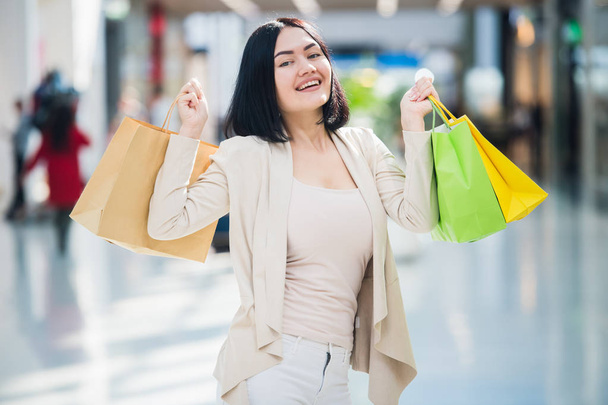 A dark-haired woman wearing muted, gentle colours holds colourful, patterned shopping bags walks at an exclusive shopping mall. - Фото, изображение