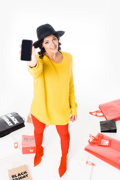 atractiva chica de moda mostrando teléfono inteligente con pantalla en blanco cerca de bolsas de compras para el viernes negro aislado en blanco
 - Foto, imagen