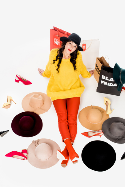 vista de ángulo alto de la joven sentada en el suelo con zapatos, sombreros y bolsas de compras con inscripción de letras de viernes negro alrededor aislado en blanco
 - Foto, imagen
