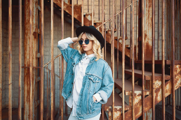 Красивая, модная молодая женщина позирует на металлической ржавой лестнице, одетая в джинсы, одежду, джинсовую куртку, белую рубашку, черную шляпу и солнцезащитные очки. Поп культура. Модный хипстерский портрет
. - Фото, изображение