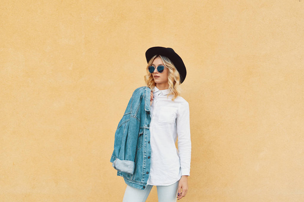 Ritratto di Hipster Fashion Girl che tiene Jaket su una spalla in piedi sullo sfondo della parete gialla. Youth Urban Fashion Concept. Copia spazio
. - Foto, immagini
