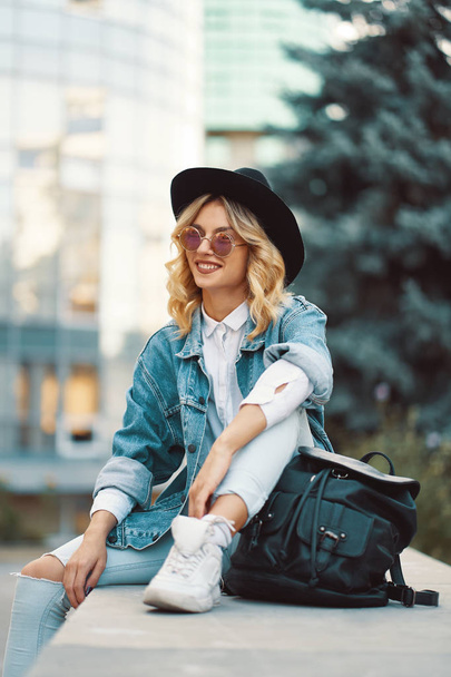 Modern életmód nő ült kint üveg épület a háttérben. Fiatal modern kaukázusi női modell visel divatos farmer, ruházat, kerek szemüveg, kalap és hátizsák cool. - Fotó, kép