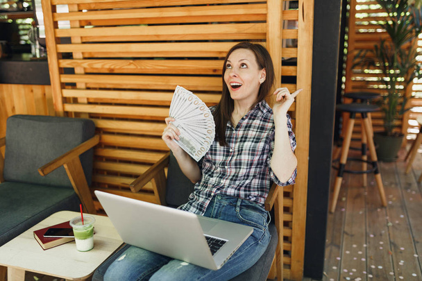 Женщина в кафе на улице сидит с современным ноутбуком, держит банкноты по доллару, наличные деньги. Мобильный офис ресторан в свободное время. Концепция внештатного бизнеса
 - Фото, изображение