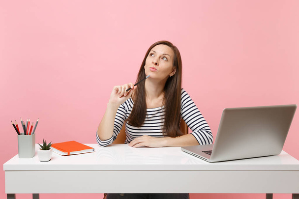 Zamyšlená žena držení tužky poblíž bradu vyhledávání si snové práce sedět na bílý stůl s moderním pc laptop izolované na pastelově růžové pozadí. Úspěch obchodní kariéru koncept. Kopírovat prostor - Fotografie, Obrázek