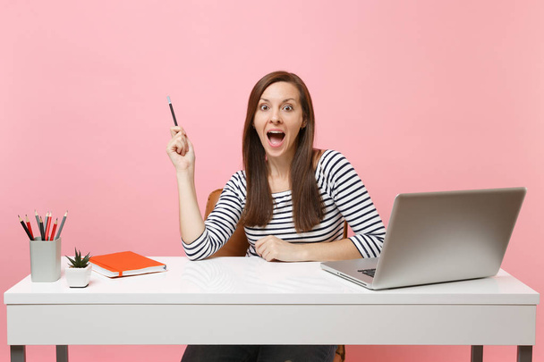 Zszokowana kobieta zaskoczony wskazującego ołówek się usiąść i pracować w biały biurko z nowoczesnym laptopem na białym tle na pastelowy różowy tło. Osiągnięcia firmy kariera koncepcja. Miejsce na reklamę - Zdjęcie, obraz