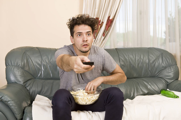 Excited young man with the remote and popcorn on the couch watching tv - Foto, imagen