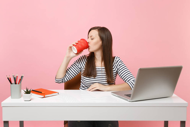 Young woman with closed eyes drink coffee or tea from cup enjoy working on project sitting at office with pc laptop isolated on pastel pink background. Achievement business career concept. Copy space - Photo, Image