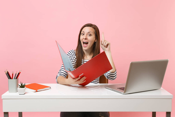 Curious woman pointing finger up holding folder with paper document work on project while sit at office with laptop isolated on pastel pink background. Achievement business career concept. Copy space - Photo, Image
