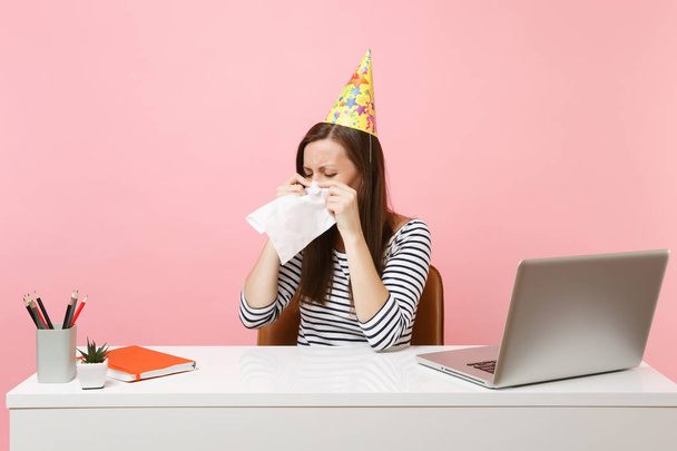 Mujer nerviosa en sombrero de fiesta llorando limpiando lágrimas con tejido porque celebrando cumpleaños solo en la oficina en el escritorio blanco con el ordenador portátil aislado en el fondo rosa. Logro de la carrera empresarial. Copiar espacio
 - Foto, imagen