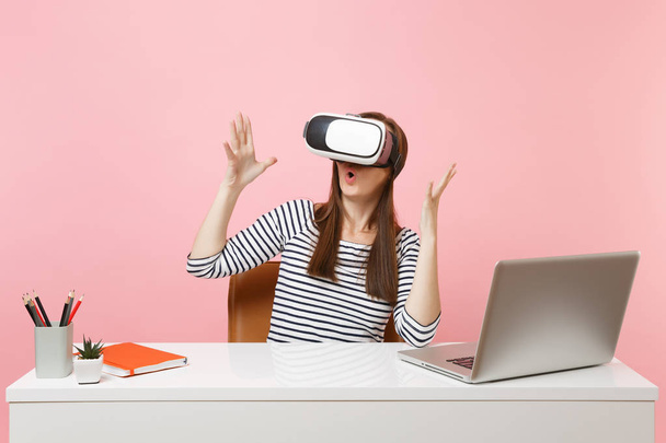 Young shocked woman in headset of virtual reality on head spreading hands sit and work at white desk with laptop isolated on pastel pink background. Achievement business career concept. Copy space - Foto, afbeelding