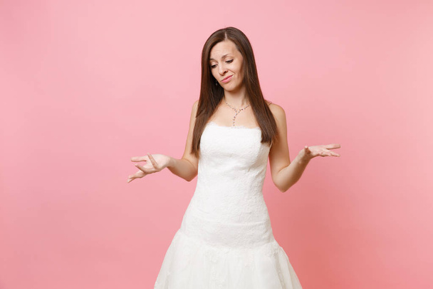 Porträt der schuldigen traurigen Braut in schönem spitzenweißen Hochzeitskleid stehend und die Hände isoliert auf pastellrosa Hintergrund ausbreitend. Hochzeitsfeier Konzept. Kopierfläche für Werbung - Foto, Bild