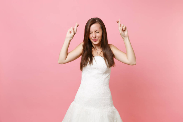 Портрет красивой невесты в белом свадебном платье, ожидающей особого момента, держащей пальцы скрещенными, глаза закрытыми изолированными на пастельно-розовом фоне. Концепция свадьбы. Копирование места для рекламы
 - Фото, изображение