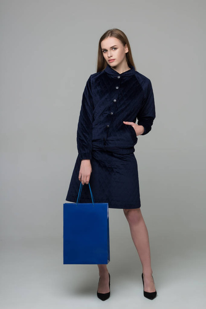 Young model long-haired blond girl in dark skirt suit stands holding blue shopping package - Zdjęcie, obraz