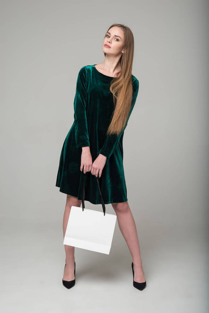 Young model long-haired blond girl in dark green short dress with lace stands holding white package with copy space - Foto, imagen