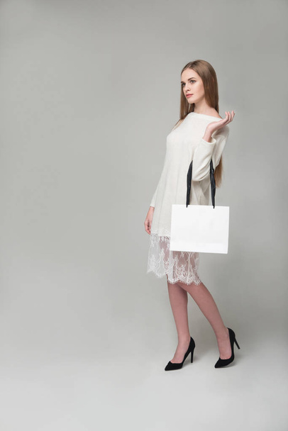 Νέος μοντέλο μακρυμάλλης ξανθό κορίτσι με λευκό κοντό φόρεμα με δαντέλες στέκεται κρατώντας λευκό πακέτο με αντίγραφο χώρου - Φωτογραφία, εικόνα