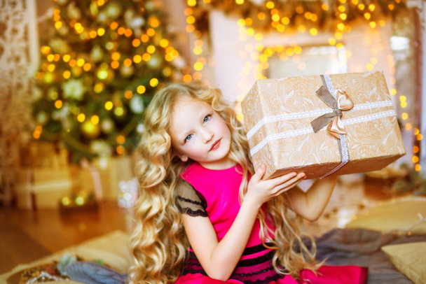 Cute little girl with long curly blond hair at home near a Christmas tree with gifts and garlands and a decorated fireplace  - Foto, afbeelding