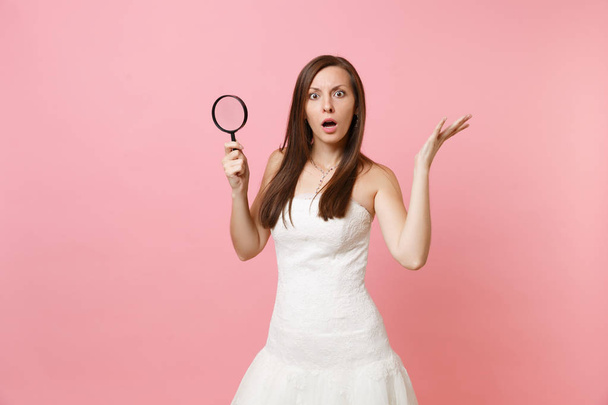 Portrait of shocked bride woman in lace white wedding dress spreading hands, holding magnifying glass isolated on pastel pink background. Wedding to do list. Organization of celebration. Copy space - Photo, image