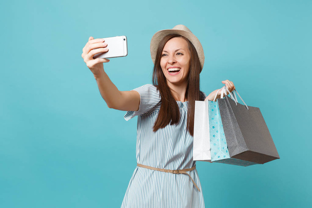 Portrait smiling woman in summer dress straw hat holding packages bags with purchases after shopping doing selfie shot on mobile phone isolated on blue pastel background. Copy space for advertisement - Foto, immagini