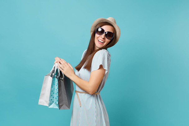 Πορτρέτο της μόδας ελκυστική ευτυχισμένη γυναίκα στο φόρεμα του καλοκαιριού, ψάθινο καπέλο, γυαλιά ηλίου, κρατώντας πακέτα σακούλες με τις αγορές μετά ψώνια απομονώνονται σε γαλάζιο παστέλ φόντο. Αντίγραφο χώρος για διαφήμιση - Φωτογραφία, εικόνα