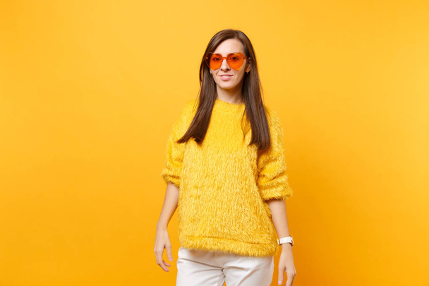 Portrait of beautiful young woman in fur sweater, white pants and heart orange glasses standing isolated on bright yellow background. People sincere emotions, lifestyle concept. Advertising area - Photo, Image