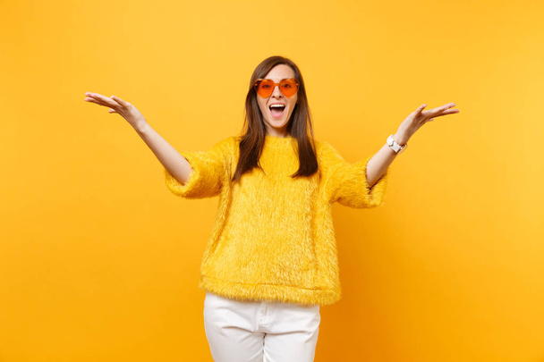 Portrait of excited happy woman in fur sweater, white pants and heart orange glasses spreading hands isolated on bright yellow background. People sincere emotions, lifestyle concept. Advertising area - Foto, Imagen