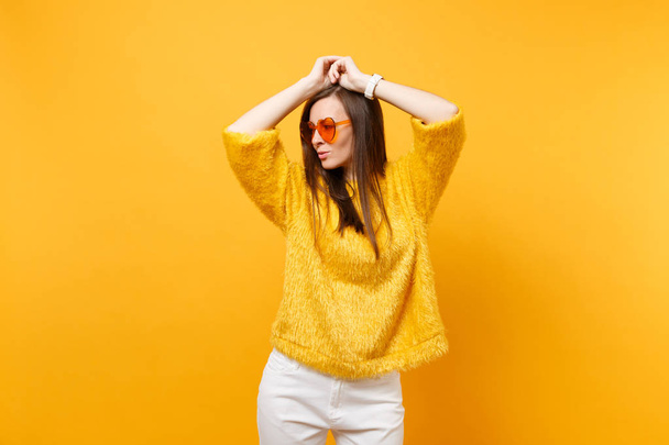 Beautiful young woman in fur sweater and heart orange glasses looking aside, putting hands on head isolated on bright yellow background. People sincere emotions, lifestyle concept. Advertising area - Photo, Image