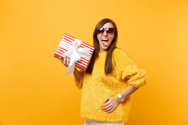 Portret van grappige jonge vrouw in rode bril tong tonen, rode doos met cadeau, huidige geïsoleerd op heldere gele achtergrond te houden. Mensen oprechte emoties, lifestyle concept. Gebied van de reclame - Foto, afbeelding