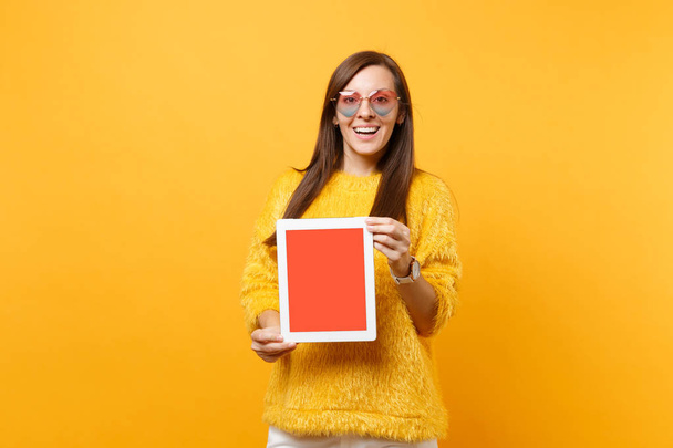 Joyful young woman in sweater, heart glasses holding tablet pc computer with blank black empty screen isolated on bright yellow background. People sincere emotions lifestyle concept. Advertising area - 写真・画像